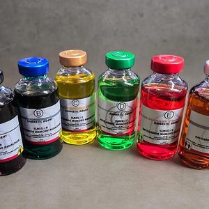 Peut inclure: Six flacons en verre remplis de liquides colorés, chacun avec une étiquette marquée des lettres A à F. Les étiquettes indiquent Amnestic Agent, Class A à F, et Retrograde Memory Reduction. Les bouchons sont noirs, bleus, bronzés, verts, rouges et argentés.