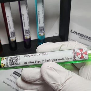 Może przedstawiać: Przezroczysta szklana probówka wypełniona jaskrawozielonym płynem, z czarną zakrętką i białą etykietą z napisem Sample // 14.386X In vitro Type // Pathogen Anti-Virus, logo Umbrella Corporation oraz kodem QR.