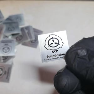 Könnte beinhalten: Eine kleine quadratische, silberfarbene Metallplatte mit gebürsteter Oberfläche, die das schwarze SCP Foundation-Logo und den Text SCP Foundation Secure. Contain. Protect. zeigt. Ein Stapel identischer Platten ist unscharf im Hintergrund.