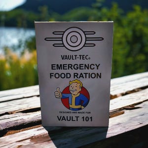 Könnte beinhalten: Eine beige Vault-Tec Notration-Verpackung. Die Verpackung zeigt das Vault-Tec-Logo, den Text "EMERGENCY FOOD RATION" und eine Zeichentrickfigur, die einen Daumen hoch zeigt. Auf der Unterseite der Verpackung steht "DESIGNED AND MADE FOR VAULT 101".
