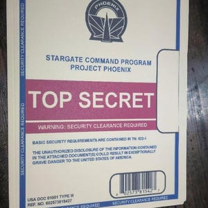 May include: A white document folder with blue borders and text reading SECURITY CLEARANCE REQUIRED. It features a Phoenix logo, the text STARGATE COMMAND PROGRAM PROJECT PHOENIX, a red bar labeled TOP SECRET, and a barcode with the number 02573 81542 7.
