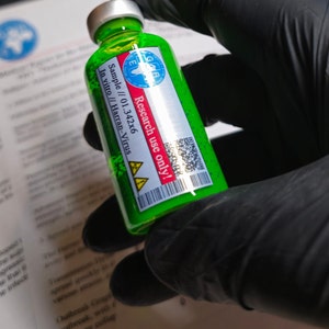 Puede incluir: Un pequeño vial de vidrio verde con una tapa plateada, lleno de un líquido verde brillante. El vial tiene una etiqueta con el texto "Research use only!" y "Harran-Virus". El vial es sostenido por una mano enguantada.
