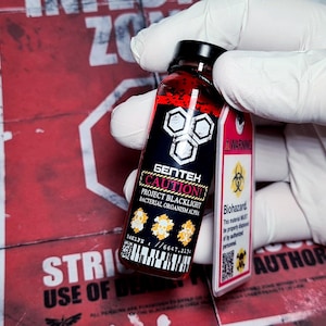 Puede incluir: Un vial con una etiqueta negra con el logotipo de GenTek y el texto "CAUTION! PROJECT BLACKLIGHT BACTERIAL ORGANISM ALPHA". Un líquido rojo llena el vial. Una etiqueta rosa con un símbolo de peligro biológico y la palabra "Biohazard" está adjunta.