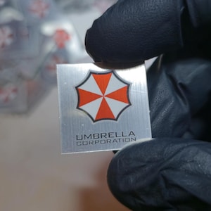 May include: A silver square featuring the Umbrella Corporation logo, a red and white umbrella design. The text "UMBRELLA CORPORATION" is printed below the logo. The item is held by a gloved hand.