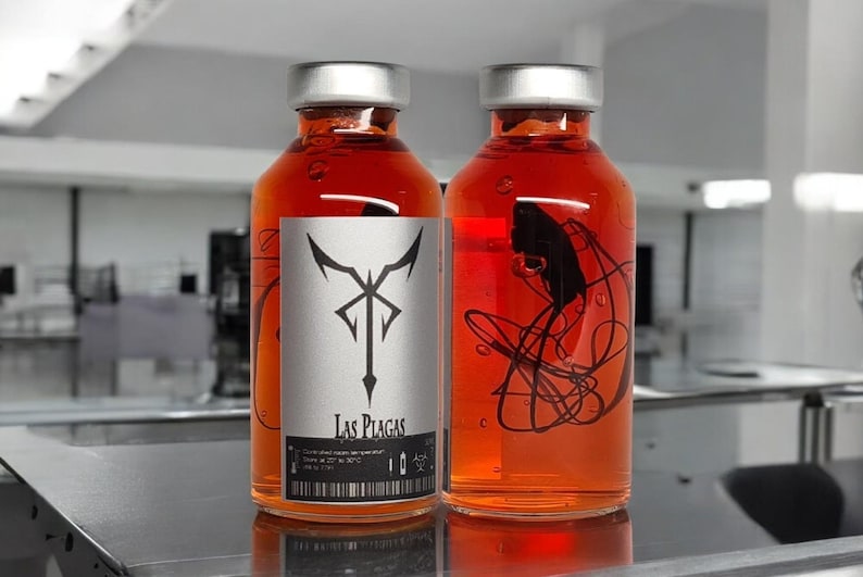 May include: Two glass bottles filled with a vibrant orange liquid, each sealed with a silver cap. One bottle features a label with a black and white design and the text "Las Plagas". The other bottle has a black abstract design inside.