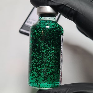 May include: A clear glass bottle filled with green liquid and black glitter. The bottle has a silver cap and a label with text. The bottle is held by a person wearing a black glove.