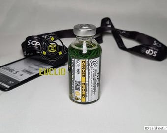 Op de afbeelding: Een doorzichtig glazen flesje gevuld met groene vloeistof, voorzien van een zilveren dop en een wit etiket met de tekst DANGER, EUCLID, SCP-199 en diverse waarschuwingssymbolen. Het flesje staat voor een zwart koord met SCP-bedrukking.