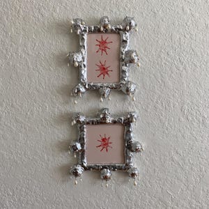 May include: Two silver-colored picture frames with a unique, textured design are mounted on a textured wall. Each frame holds a pink background with a red starburst design. The frames are adorned with pearl-like embellishments.