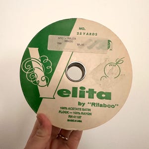 May include: A spool of green and white ribbon with the text "Velita by "Rifabco" 100% Acetate Satin Flock - 100% Rayon RN 41137 Made in USA" printed on it. The spool is labeled "No. 25 Yards" and "$8.00".