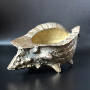 May include: A gold-colored seashell planter with a textured surface and a wide opening. The planter has three legs and is made of metal.