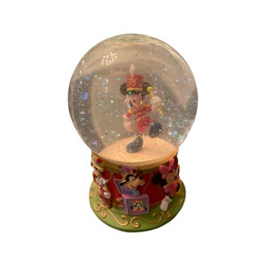 May include: A Disney snow globe featuring Mickey Mouse in a red and gold band uniform, holding a baton. The base is decorated with Disney characters. The globe is filled with glitter and has a clear glass dome. The number 75 is on Mickey's hat.