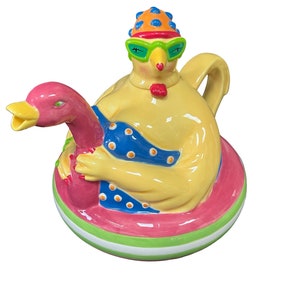 Puede incluir: Una caprichosa tetera de cerámica con forma de pollo amarillo con gafas de sol y gorro de baño, abrazando a un pato rosa sobre un flotador rosa, azul y verde. La tetera tiene un asa y un pico.