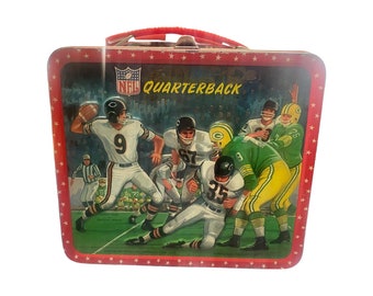 May include: Vintage metal lunchbox featuring an NFL football game scene with the word "Quarterback". The lunchbox has a red handle and a red border with white stars. The image depicts players in white and green uniforms.