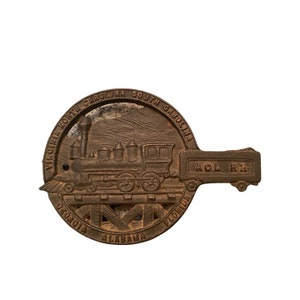 May include: A vintage bronze-colored railroad key featuring a detailed steam train design. The key is circular with a handle and includes the text "Virginia North Carolina South Carolina Georgia Florida Alabama" and "ACL RR".