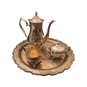 Könnte beinhalten: Ein silbernes Teeservice auf einem dekorativen Tablett. Das Set umfasst eine Teekanne mit Deckel, einen Sahnekännchen und eine Zuckerdose mit Deckel. Das Tablett hat einen gewellten Rand. Das Set ist aus glänzendem Silbermetall gefertigt.