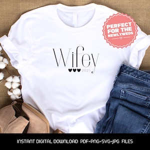 Puede incluir: Camiseta blanca con la palabra "Wifey" en escritura negra, tres corazones negros y el año "2025". Una pegatina rosa con el texto "Perfect for the Newlyweds" es visible. La camiseta se combina con unos vaqueros azules.