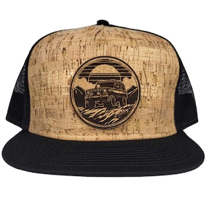 May include: A brown cork trucker hat with a black mesh back. The hat features a round, wood-burned patch with a silhouette of a jeep driving through a mountain landscape.