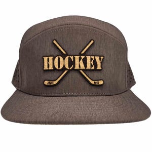 以下が含まれることがあります： グレーとブラックのベースボールキャップ。ブラウンのパッチには「HOCKEY」の文字と交差した2本のホッケーのスティックが描かれている。