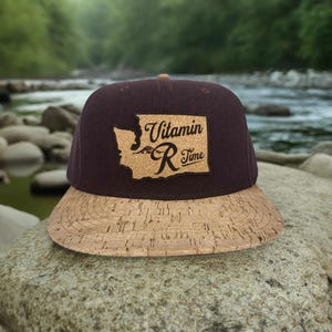 May include: A brown and tan baseball cap with a cork brim. The front of the cap features a brown patch with the outline of the state of Washington and the text "Vitamin R Time".