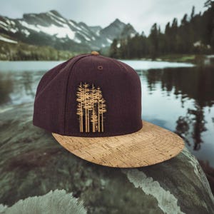 May include: A burgundy and cork baseball cap with a brown silhouette of a group of trees on the front.