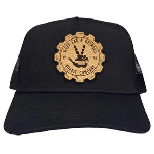 May include: Black trucker hat with a brown cork patch featuring a gear with the text "Sleep Eat & Offroad Est. 1986 Korkit Company" and a peace sign with a Jeep silhouette.