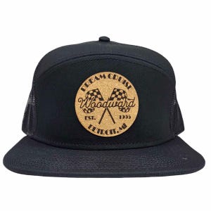 May include: Black trucker hat with a flat brim and mesh back. The front features a round, cork-like patch with the words "Dream Cruise Woodward" and crossed checkered flags. The text "Detroit, MI" and the year "1995" are also visible.