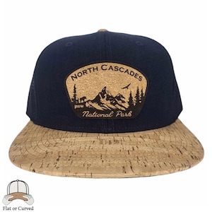 May include: A navy blue and cork baseball cap with a cork patch featuring a mountain scene and the text "North Cascades National Park".