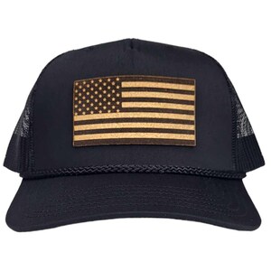 Pode incluir: Um boné de caminhoneiro preto com um remendo de couro marrom da bandeira americana na frente.
