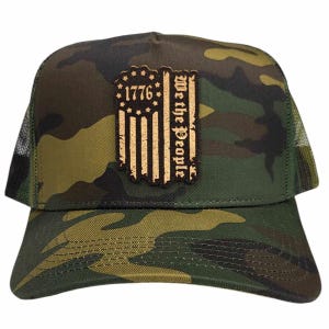 Pode incluir: Boné de caminhoneiro camuflado com um remendo de couro marrom apresentando uma bandeira americana estilizada com o texto "1776 We the People".