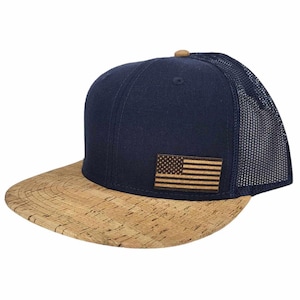 Pode incluir: Um boné de beisebol azul marinho e marrom com uma aba de cortiça e um pequeno patch da bandeira americana na frente.