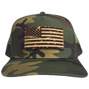 Pode incluir: Um boné de caminhoneiro camuflado com um remendo de bandeira americana desgastado. A bandeira é feita de madeira e tem uma mancha marrom.