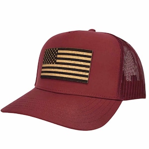 Pode incluir: Um boné de caminhoneiro bordô com um remendo de couro marrom com a bandeira americana na frente. O boné tem uma parte de trás de malha.