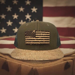 Pode incluir: Um boné de caminhoneiro verde e preto com uma aba de cortiça. O boné apresenta um remendo de bandeira americana desbotado feito de cortiça.