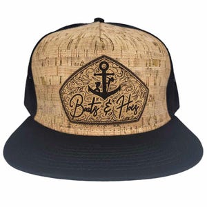 Pode incluir: Um boné de caminhoneiro de cortiça marrom e malha preta com um remendo de couro preto com uma âncora e o texto "Boats & Hoes".