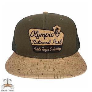 May include: Olive green and cork baseball cap with a mesh back. The front features a cork patch with the text "Olympic National Park Puddle Jump'n & Raindogs" and a forest graphic. The brim is made of cork.