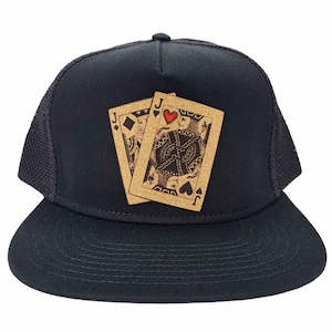 Op de afbeelding: Een zwarte trucker-pet met een bruin en gouden ontwerp van twee speelkaarten, de boer van harten en de boer van schoppen.