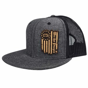 Pode incluir: Um boné de caminhoneiro preto e cinza com um remendo de bandeira americana desbotada que diz "1776 We the People".