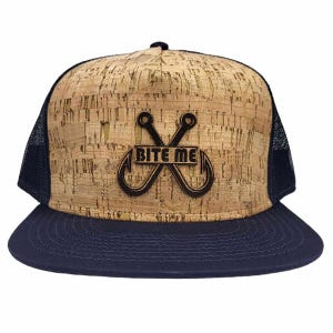 May include: A trucker hat featuring a natural tan cork front panel with a dark brown graphic of two crossed fishing hooks and the text BITE ME in the center. The hat has a solid navy blue flat brim and navy blue mesh side and back panels.