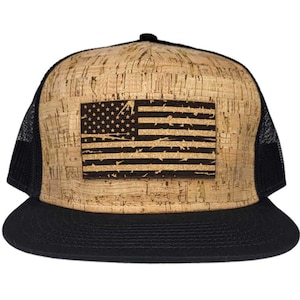 Pode incluir: Um boné de caminhoneiro preto e marrom com um exterior de tecido de cortiça e uma parte de trás de malha preta. O boné apresenta um remendo de bandeira americana desbotado na frente.