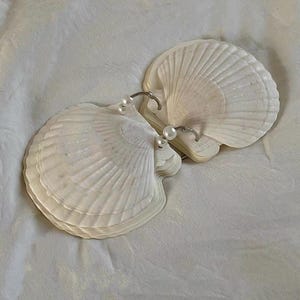 May include: A pair of white seashell earrings with small, round, white pearls. The earrings are set on silver hoops and rest on a white, textured surface. The shells have a fan-like design.