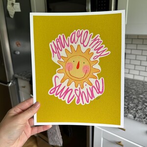 May include: A watercolor print on yellow paper with the words "you are my sunshine" in pink script. The image features a smiling sun with pink cheeks and a red nose. The print is held by a person.