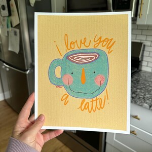 May include: A whimsical art print featuring a teal coffee cup with a smiling face, orange nose, and pink cheeks. The words "i love you a latte!" are written in orange script on a yellow background.