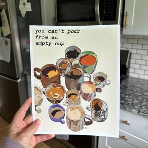 May include: A print featuring various coffee cups with different coffee and cream designs. The text "you can't pour from an empty cup" is displayed at the top. The artwork is held by a person in a kitchen setting.
