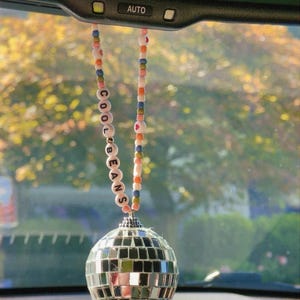 Può includere: Un ornamento per specchietto retrovisore per auto con un design a sfera da discoteca. L'ornamento è appeso a una catena di perline con le parole "COOL BEANS" scritte con perline bianche. Lo sfondo mostra una vista sfocata di alberi e l'ora e la data.