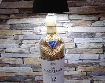 Può includere: Una lampada a forma di bottiglia di vetro trasparente con un paralume nero. La bottiglia è piena di orzo e ha un'etichetta che recita "The Macallan Highland Single Malt Scotch Whisky 12 Double Cask".