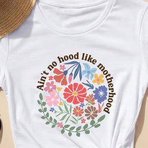 Peut inclure: T-shirt blanc avec un motif floral et le texte "Ain't no hood like motherhood" dispos&eacute; en cercle. Le motif pr&eacute;sente des fleurs color&eacute;es et des feuilles vertes. Le t-shirt est nou&eacute; &agrave; la taille.