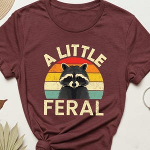 May include: Burgundy t-shirt with a raccoon graphic inside a retro sunset design. The text "A LITTLE FERAL" is above and below the raccoon. Accessories include sunglasses, a watch, and a bracelet.