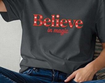 May include: Dark grey heather t-shirt with the words "Believe in magic" in red with white snowflake accents. The shirt is a crew neck style and is made of a soft material. The model is wearing blue jeans.