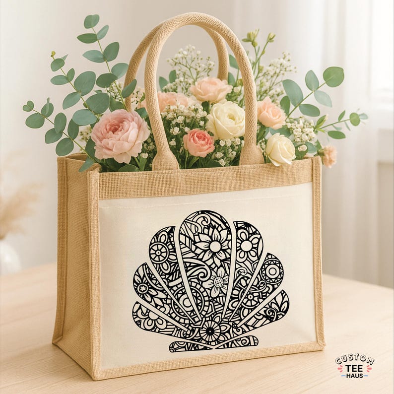 May include: A beige jute tote bag with a white front panel featuring a black seashell design filled with floral patterns. The bag has beige handles and is filled with pink and white roses and greenery.