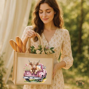May include: A beige tote bag with a watercolor illustration of a Highland cow in a pink pickup truck. The bag also contains baguettes and flowers. The words "CUSTOM TEE HAUS" are in the lower right corner.